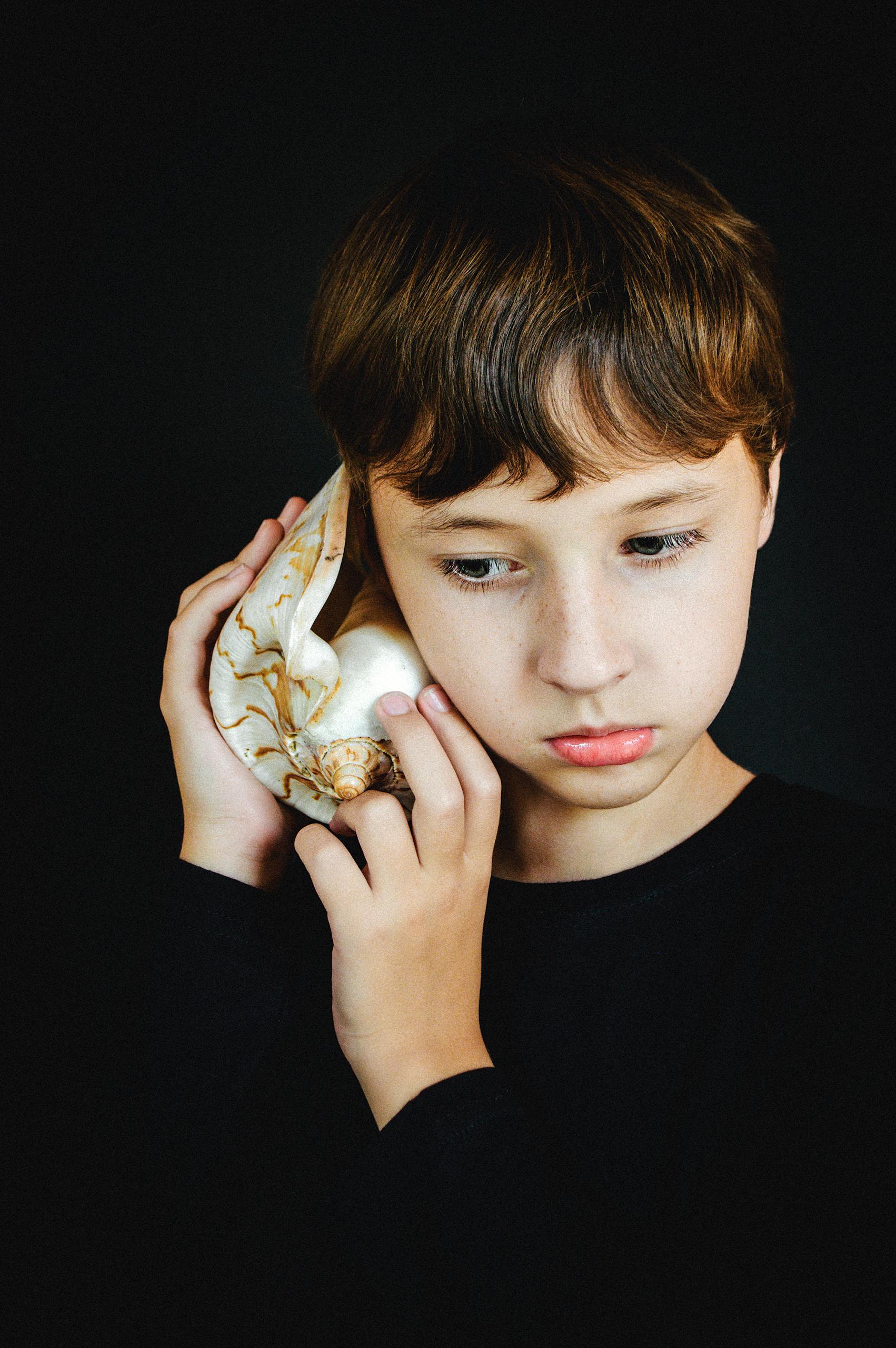 A young boy with brown hair listens to a large seashell against a black background.